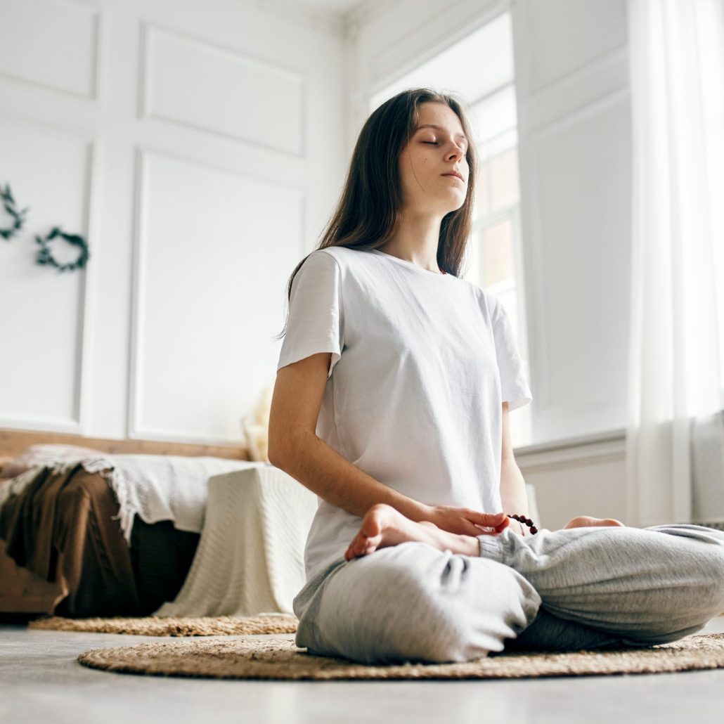 Full length of woman practicing breathing exercise. Young woman with eyes closed sitting in lotus position. She is living room at home.